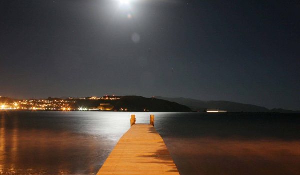Looking down a dock in the nighttime at the ocean and Wellington shoreline in the distance.